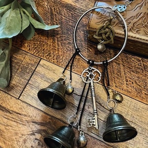 May include: A silver metal wind chime featuring a large ring, black cords, and various charms. The charms include three bronze bells, an ornate key, and small decorative bells. A Celtic knot charm is also present.