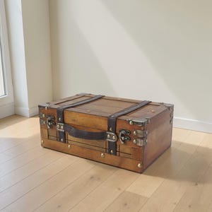 Op de afbeelding: Een houten koffer in vintage-stijl met metalen hoekbeschermers en leren riemen. De koffer heeft een donkerbruine leren handgreep en messingkleurige hardware. De koffer is rechthoekig en staat op een lichte houten vloer.