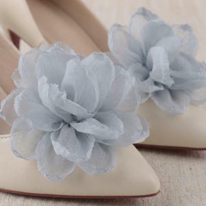 May include: A pair of cream-colored pointed-toe shoes, each adorned with a large, layered, light blue fabric flower on the toe. The flowers feature delicate, semi-sheer petals with slightly ruffled edges, creating a voluminous, textured appearance.