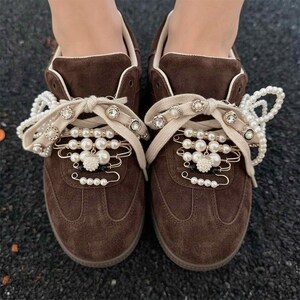 Op de afbeelding: Een paar donkerbruine suède sneakers met beige veters versierd met meerdere zilverkleurige veiligheidsspelden, witte kunstparels en kleine doorzichtige strass-steentjes. De decoratieve spelden zijn in rijen op de tong geplaatst.