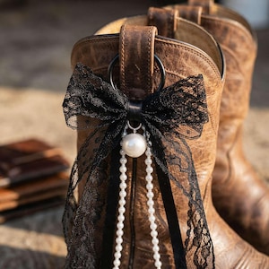 May include: A brown leather cowboy boot featuring a decorative accessory attached to the pull tab. The accessory consists of a black lace bow, a single large white faux pearl, and two long strands of small white faux pearls hanging downward.