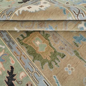 May include: A woven area rug featuring a complex pattern with a tan background and accents in light blue, sage green, cream, and dark brown. The design includes geometric and floral-inspired motifs arranged in a repeating layout with borders.