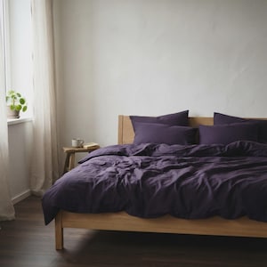 Op de afbeelding: Een houten bedframe met een dieppaarse dekbedovertrek en bijpassende kussenslopen. Het beddengoed heeft een zachte, licht gekreukte textuur en het bed staat tegen een effen witte muur, wat zorgt voor een eenvoudige en strakke slaapkamerinrichting.