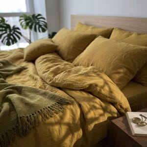 May include: A mustard yellow bedding set featuring a duvet cover and multiple pillows, accompanied by a textured olive green throw blanket with fringe edges. The fabric has a visible linen-like weave, creating a soft and layered appearance.