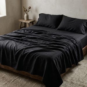 Può includere: Un set di biancheria da letto in lino nero antracite composto da un lenzuolo, un lenzuolo con angoli e due federe abbinate. Il tessuto presenta una trama visibile ed è drappeggiato su una struttura letto in legno. La biancheria è scura e opaca.