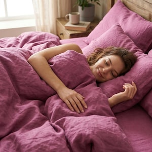 Puede incluir: Una persona durmiendo en una cama con ropa de cama de color malva. La ropa de cama incluye una funda nórdica, almohadas y sábanas. La cama está cerca de una ventana y una mesita de noche de madera con una taza y una planta.