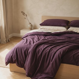 May include: A bed featuring a deep plum-colored duvet cover and matching pillowcases layered over cream-colored sheets. The bed has a light-toned wooden frame and headboard, with a small vase containing a dried plant branch placed nearby.