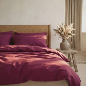 May include: A deep magenta duvet cover and matching pillowcases on a wooden bed frame. Beside the bed, a light-colored ceramic vase holds a large arrangement of dried grasses and flowers, resting on a small, rustic wooden stool.