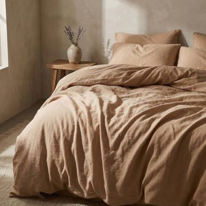 May include: A bed with tan-colored bedding, including a duvet cover and pillows. A small wooden side table holds a vase with lavender. The room has a neutral color palette, creating a calming and inviting atmosphere.