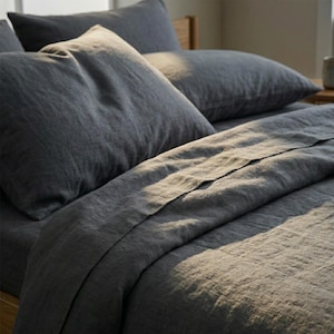 May include: A bed dressed with dark grey linen bedding. The image shows a fitted sheet, a flat sheet, and several pillows. The fabric has a textured appearance, and the lighting creates shadows and highlights.