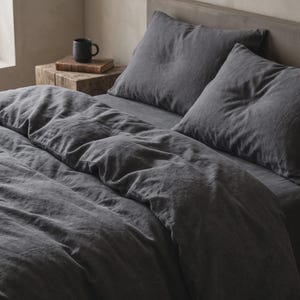 May include: A charcoal grey bedding set featuring a duvet cover and two matching pillows with a visible textured fabric weave. The bedding is arranged on a bed frame with a headboard, accompanied by a dark mug resting on a wooden bedside table.