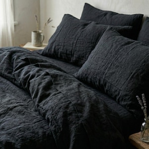 Puede incluir: Juego de ropa de cama de lino de color carbón oscuro, que incluye una funda nórdica y varias fundas de almohada. La tela tiene un aspecto texturizado y arrugado. La ropa de cama está colocada sobre una cama, creando un ambiente acogedor. Tema: decoración del hogar.