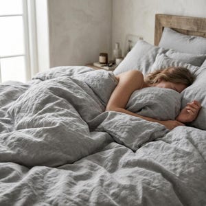 May include: A bed with grey linen bedding, including a duvet cover and pillows. A person is sleeping, partially covered by the bedding. The wooden headboard is visible in the background. The room has a neutral colour palette.