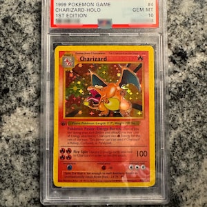 May include: A graded 1999 Pokémon Game Charizard-Holo 1st Edition card. The card features a vibrant illustration of Charizard, a fire-type Pokémon. The card is encased in a protective case with a GEM MT 10 grade. The text on the card includes the Pokémon's name, stage, and attack details.
