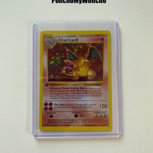May include: A holographic Charizard trading card in a clear plastic protective sleeve. The card features a yellow border, the name Charizard, 120 HP, and an illustration of a fire-breathing dragon. Text includes Pokémon Power Energy Burn and Fire Spin.