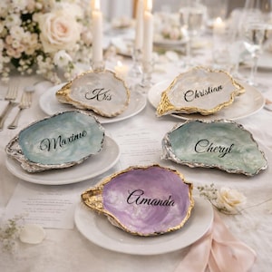 Pode incluir: Cinco cartões de lugar decorativos em forma de conchas de ostra com nomes escritos em caligrafia elegante. Cada concha tem uma cor única, incluindo branco, azul e roxo, com bordas douradas ou prateadas. As conchas estão dispostas em pratos brancos.