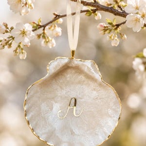 Op de afbeelding: Een schelpvormig ornament in gebroken wit met een goudkleurige rand en de gouden letter "A". Het hangt aan een crèmekleurig lint tegen een vage achtergrond van bloeiende takken. Een decoratief item.