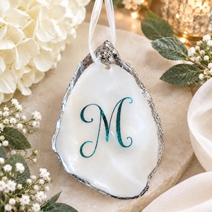 Könnte beinhalten: Ein weißes Austernschalen-Ornament mit einem türkisfarbenen Glitzerbuchstaben M in der Mitte. Die Schale hat einen metallisch silber bemalten Rand und ist an einer dünnen weißen Bandschlaufe befestigt, ein dekoratives Stück zum Aufhängen.