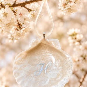 May include: A seashell-shaped ornament with a cream-colored, marbled appearance, adorned with a silver-colored letter "H". The ornament hangs from a cream-colored ribbon against a backdrop of white blossoms.