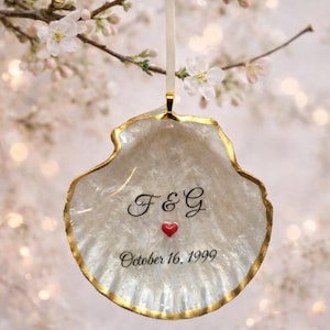 May include: A pearlescent, shell-shaped ornament with a gold-painted edge, hanging from a white ribbon. The ornament features the initials "F&G", a red heart, and the date "October 16, 1999". The background is a soft, blurred floral arrangement.