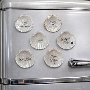 Op de afbeelding: Een verzameling schelpvormige magneten met verschillende opschriften. De magneten zijn bevestigd op een zilveren koelkast. Tekst omvat "Baby", "F&G", "75494", "Be Kind", "Love, Mom" en "But first, Coffee".