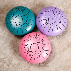 May include: Three colorful steel tongue drums in teal, lavender, and pink. Each drum has a circular shape with cut-out tongues and numbers printed in gold. The drums are arranged on a neutral-colored surface.