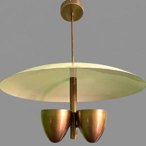 May include: A mid-century modern pendant light fixture. It features a large, off-white, saucer-shaped shade and two copper-colored, cup-shaped lights. The fixture is suspended from a brass-colored rod and ceiling mount.