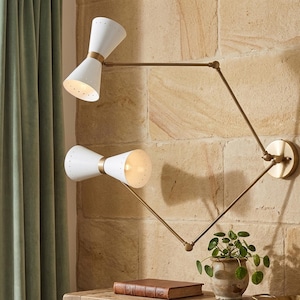 Op de afbeelding: Een witte, dubbelkegelvormige wandlamp met messing accenten en een messing arm. De lamp is bevestigd aan een getextureerde stenen muur. De lamp heeft twee lampen en het licht is aan. Een kleine potplant staat eronder.