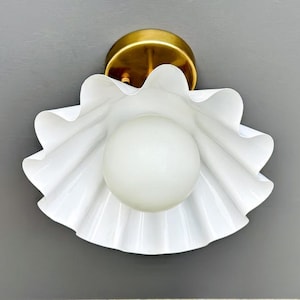 May include: A white, ruffled-edge wall sconce with a frosted globe bulb. The sconce has a brass-coloured base and is mounted on a grey wall. The design is reminiscent of a flower.