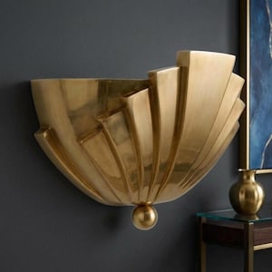 May include: A gold-coloured, Art Deco-style wall sconce. The sconce has a fan-shaped design with stepped elements and a rounded base. It is mounted on a dark grey wall.
