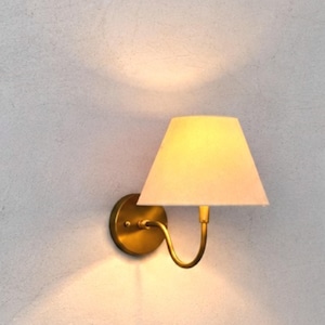 May include: A wall-mounted light fitting featuring a circular brass base, a curved brass arm, and a tapered off-white fabric lampshade. The lamp emits a warm, soft glow that illuminates the area above and below the shade, creating light patterns.
