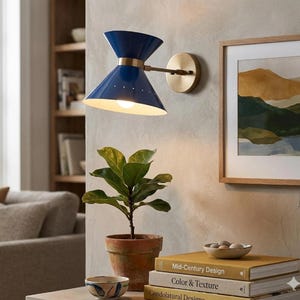 May include: A blue and brass wall sconce with a conical shade and a round backplate. The sconce is mounted on a wall next to a framed landscape painting. A potted plant and books are on a table below.
