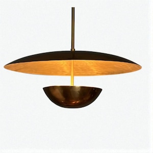 Op de afbeelding: Een moderne hanglamp met een zwarte, schotelvormige kap en een goudkleurig interieur. Daaronder bevindt zich een kleiner, komvormig element. De lamp hangt aan een messingkleurige stang, wat zorgt voor een stijlvol, eigentijds ontwerp.