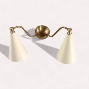 May include: A wall-mounted light fixture with two cream-colored, cone-shaped shades. The fixture has a brass finish, with curved arms extending from a central sphere. The design is minimalist and modern, suitable for interior lighting.