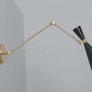 Może przedstawiać: Nowoczesna lampa ścienna z mosiężnym wykończeniem i czarnym stożkowym kloszem. Lampa posiada regulowane ramię i okrągłą podstawę, idealną do czytania lub oświetlenia akcentującego. Klosz ma złote wnętrze.