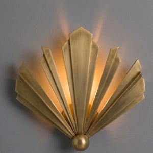 May include: A brass wall sconce with a fan-shaped design. The sconce features vertical, elongated panels that radiate from a central point. A small, round sphere is at the base. Light shines from behind the panels, creating a warm glow.