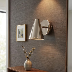Op de afbeelding: Een gepolijste zilveren wandlamp met een conische kap en een ronde achterplaat. De lamp is bevestigd aan een getextureerde, donkergrijze muur. Een kleine ingelijste foto en een vaas met groen zijn ook zichtbaar.