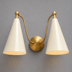 Può includere: Una lampada da parete doppia con due paralumi conici color bianco sporco fissati a bracci in ottone curvi. L'apparecchio include una piastra di montaggio centrale circolare in ottone che collega entrambi i bracci, creando un design simmetrico.