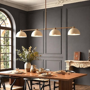 May include: A linear chandelier featuring four matte cream-colored dome shades suspended from a horizontal brass rod. The fixture is mounted to the ceiling with a central vertical stem, providing downward illumination over a dining table.