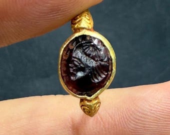 May include: A gold pendant featuring a dark red, oval gemstone with an intricate carved design. The pendant is held between fingers against a dark backdrop. The gold setting has decorative detailing. The pendant is likely an antique.