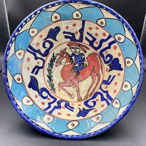 Puede incluir: Cuenco de cerámica pintado a mano con una ilustración central de una persona montando un caballo rojo. El cuenco presenta un borde azul y blanco con patrones decorativos y texto. La paleta de colores incluye rojo, azul y crema.