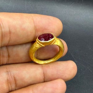Op de afbeelding: Een gouden ring met een grote, ovaalvormige, rode edelsteen. De ring heeft een dikke band en een textuurontwerp rond de edelsteen setting. De ring wordt in een hand gehouden tegen een donkere achtergrond.