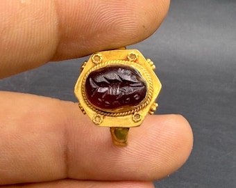 May include: A gold ring featuring a dark red, oval-shaped stone set within a hexagonal frame. The ring is held between two fingers. The stone has an engraved design. The ring's band and setting are detailed with small decorative elements.