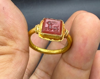 May include: A gold ring with a red, rectangular gemstone, engraved with an animal. The ring has a twisted rope design around the gemstone and a plain gold band. The ring is held in a hand against a dark background.