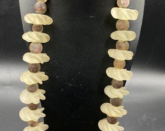May include: A necklace featuring alternating light-colored, carved, oval-shaped beads and dark, round beads. The necklace is displayed on a black stand, showcasing its unique design and color contrast. The beads are strung on a gold-colored cord.