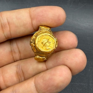 Peut inclure: Une petite bague antique de couleur or avec un design détaillé. La bague présente un emblème circulaire avec une figure centrale et des motifs complexes. La bague est tenue dans une main sur un fond sombre.