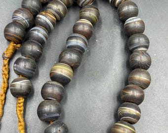 May include: A necklace of dark brown, round beads with white and tan stripes. The beads are strung on a gold-colored cord. The beads have a polished appearance and vary slightly in size. The necklace is displayed against a dark background.