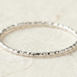 May include: A delicate silver ring with a textured surface. The ring has a thin band with a repeating geometric pattern. The ring is shown on a light-colored, textured background.