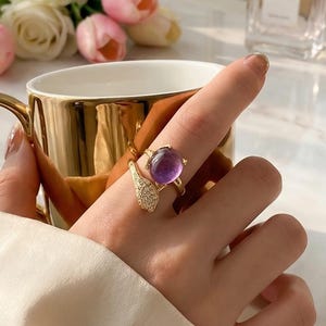 May include: A gold ring featuring a large, oval, purple gemstone and a decorative, wing-shaped design. The ring is adorned with small, clear crystals. The ring is on a finger, with a gold cup and flowers in the background.