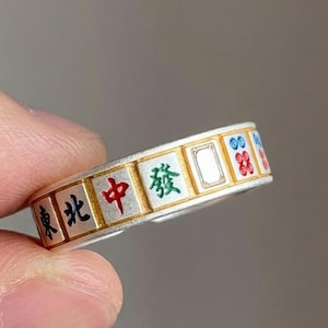May include: A silver ring featuring a repeating pattern of colourful Mahjong tiles. The tiles display Chinese characters in red, green, and blue. The ring is held between fingers against a blurred background.